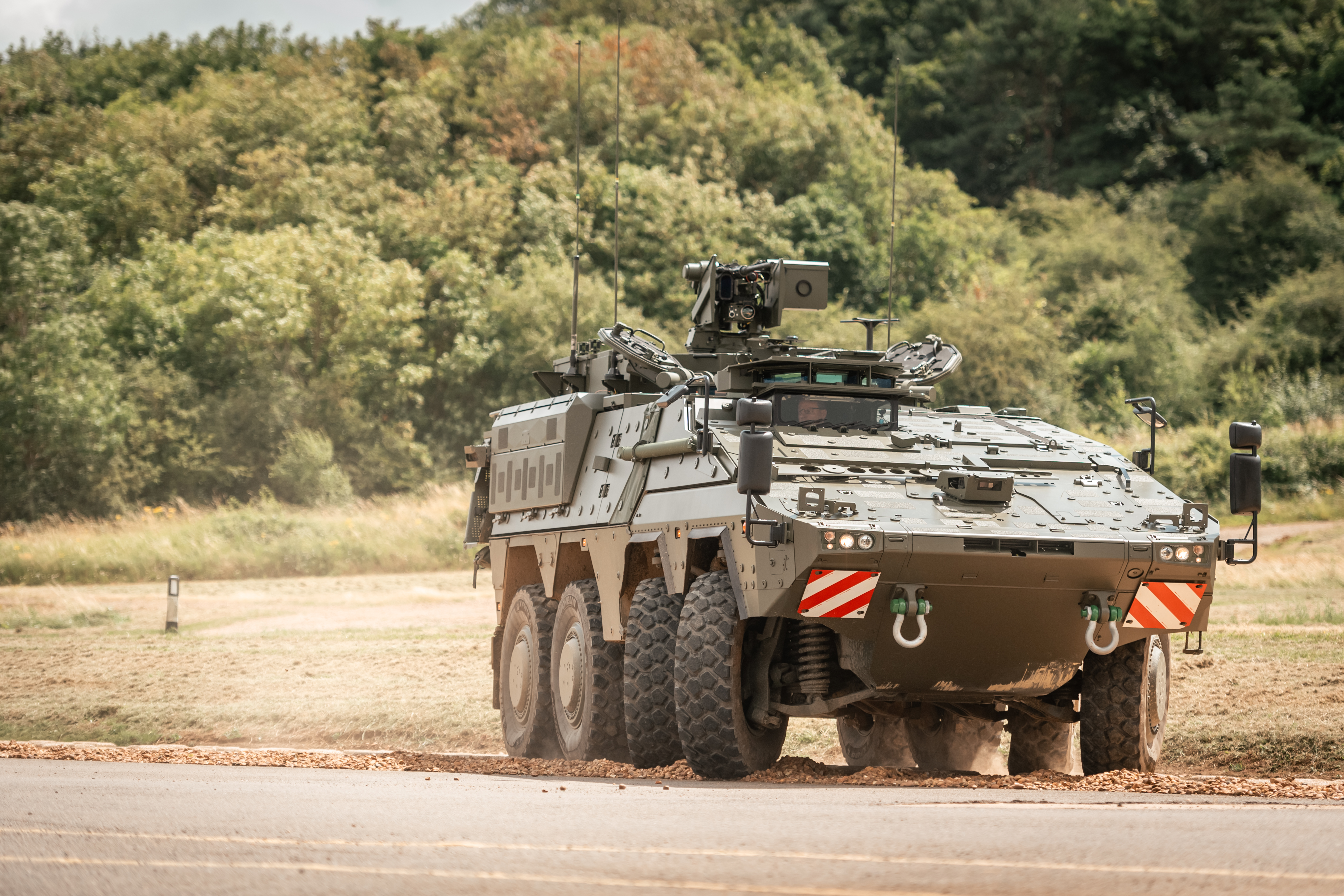 A military vehicle, specifically a Boxer, is positioned on a dirt path near a grassy expanse with dense trees.