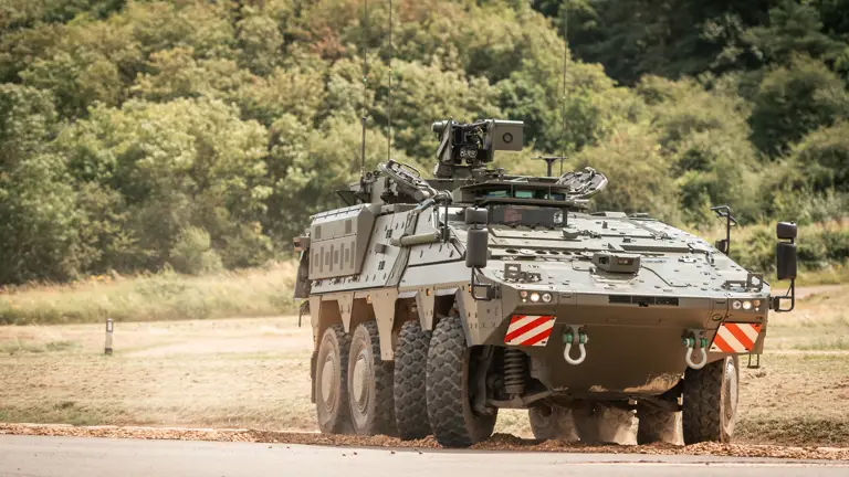 A military vehicle, specifically a Boxer, is positioned on a dirt path near a grassy expanse with dense trees.