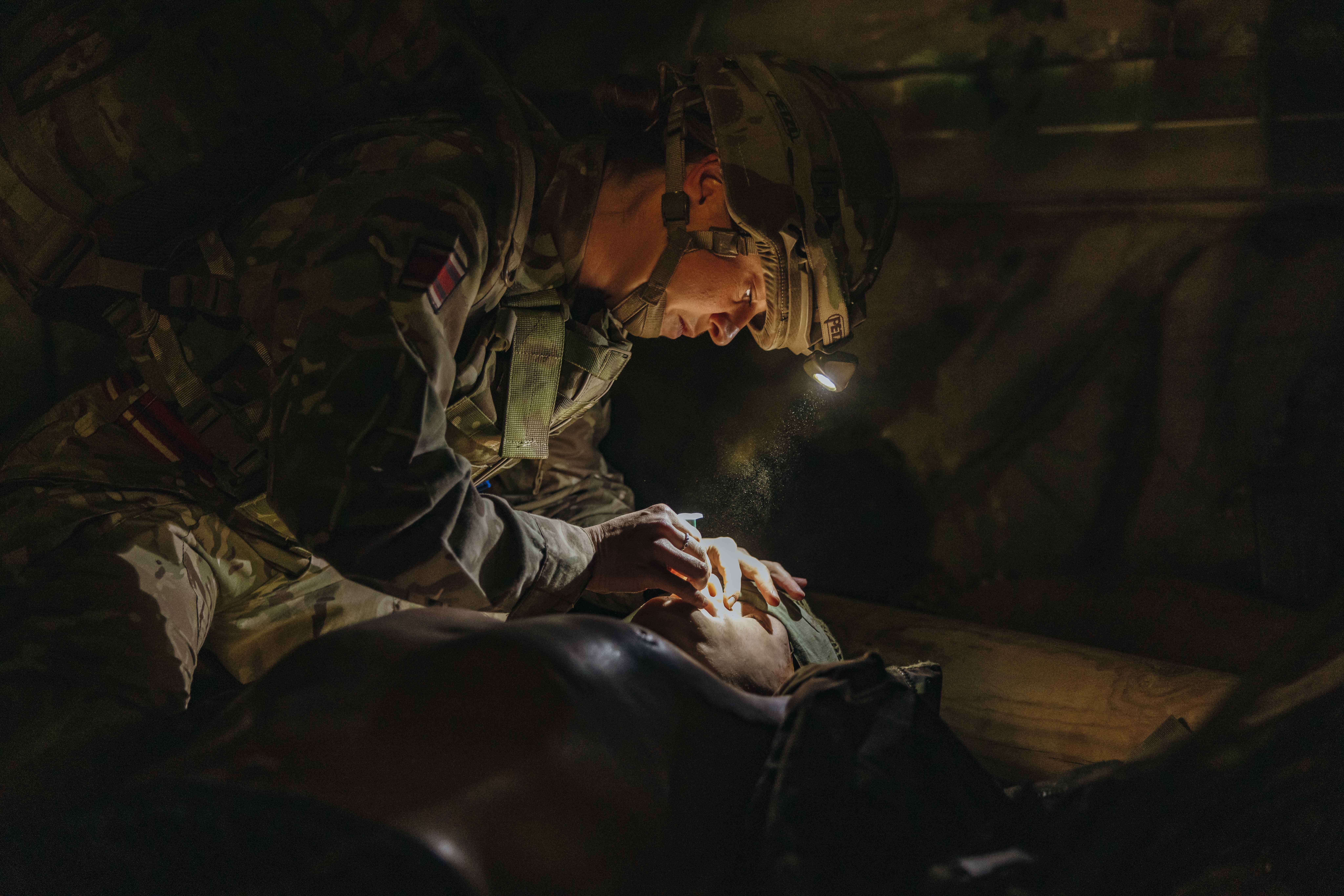Soldier wearing camouflage and helmet with a headlamp providing medical aid in a dimly lit environment.