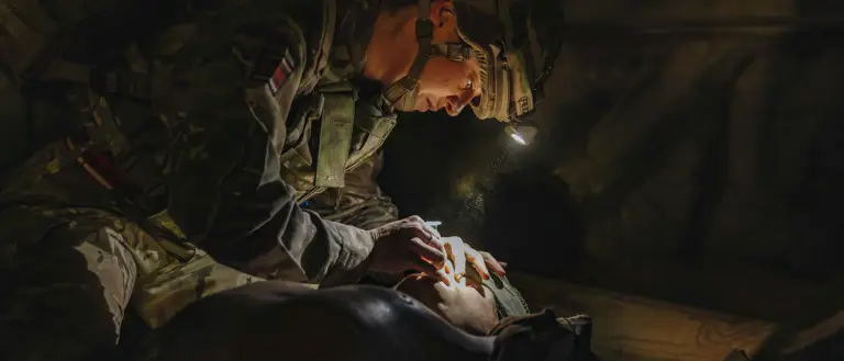 Soldier wearing camouflage and helmet with a headlamp providing medical aid in a dimly lit environment.