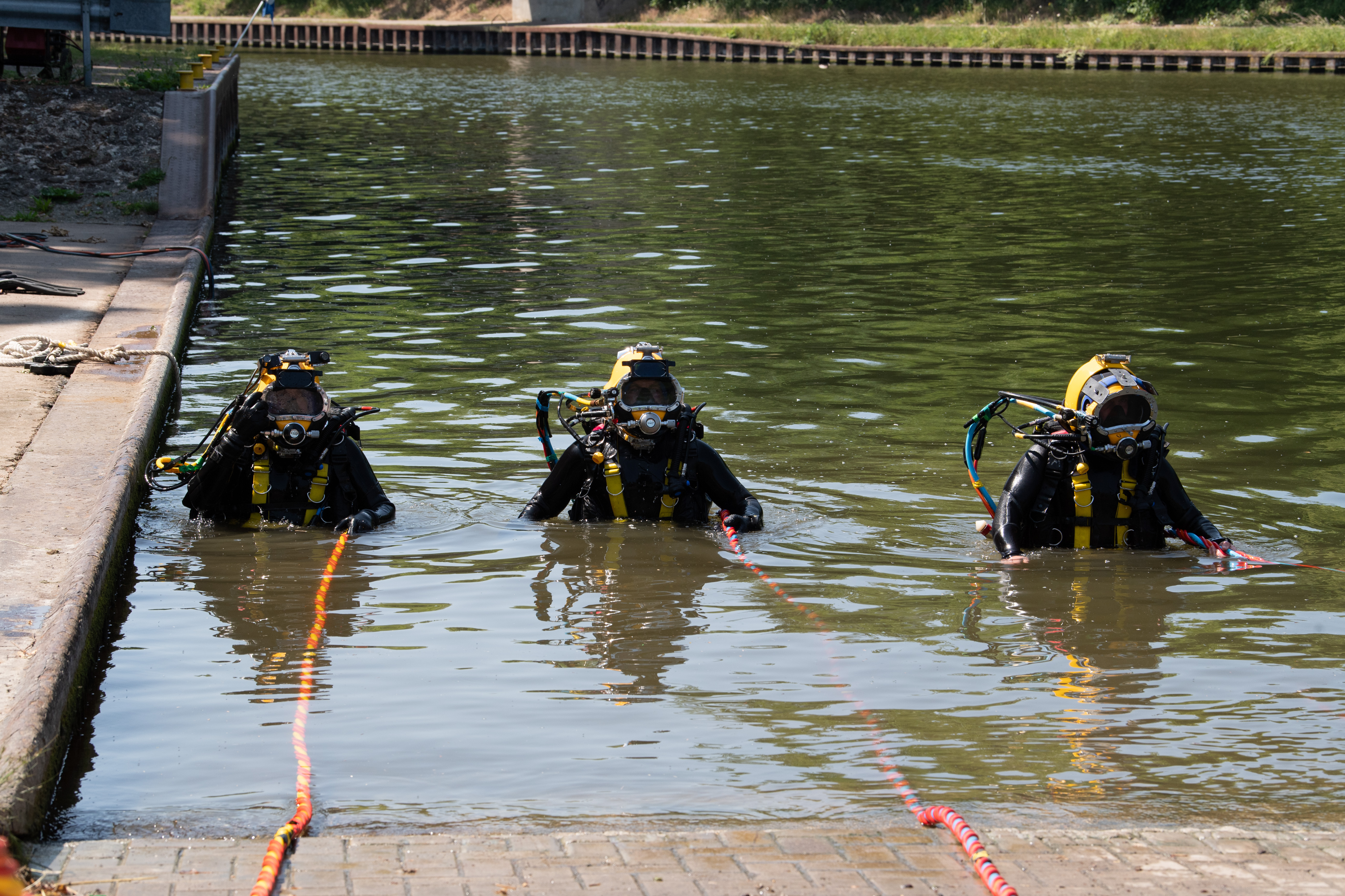 Three divers in full gear with yellow helmets stand waist-deep in a calm water canal near a concrete dock.