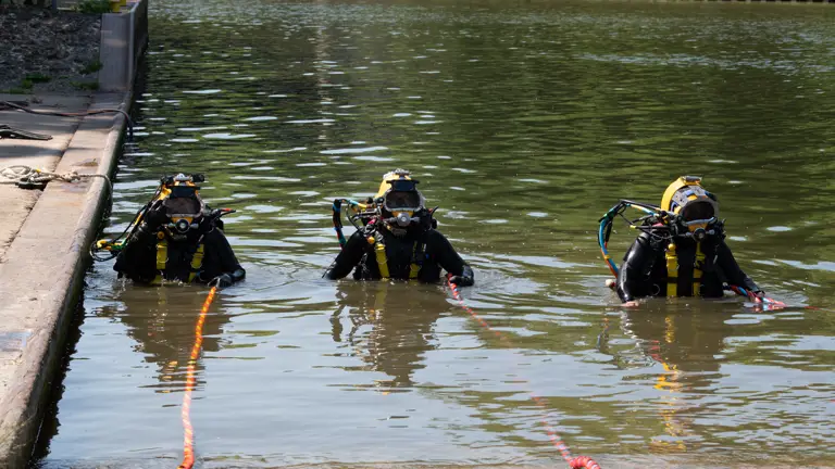 Three divers in full gear with yellow helmets stand waist-deep in a calm water canal near a concrete dock.