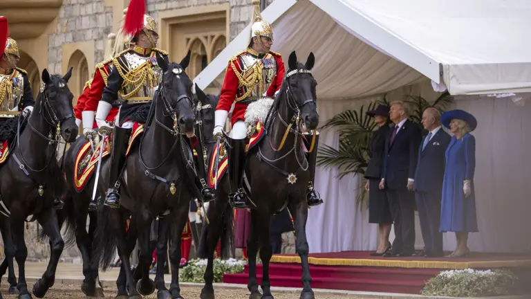Mounted guards in ornate red and gold uniforms ride black horses past dignitaries standing on a red-carpeted platform.