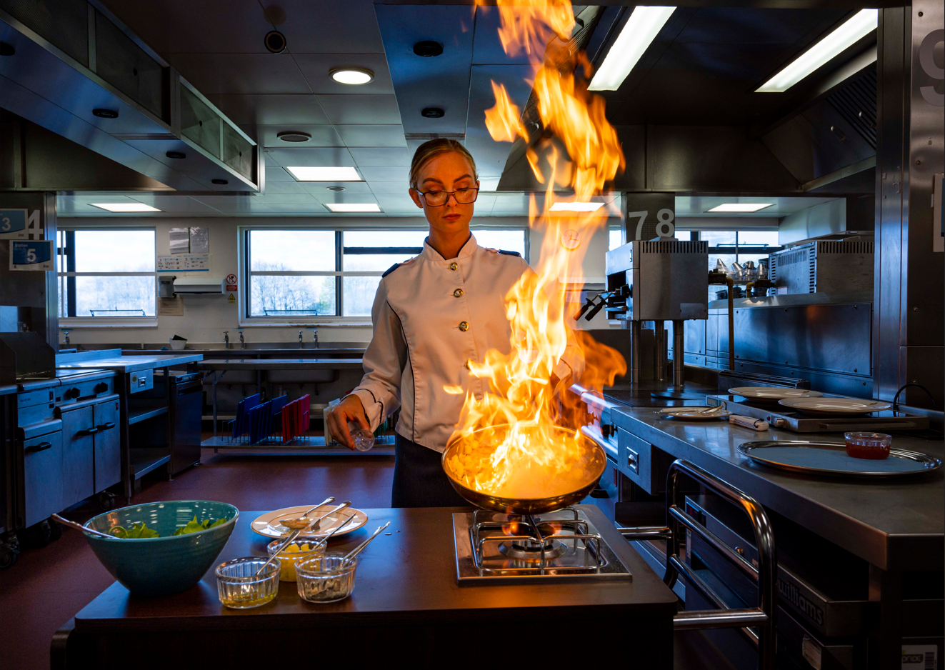 Chef flambéing a dish in a pan with tall flames in a professional kitchen setting.