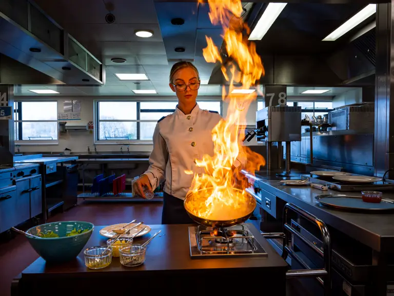 Chef flambéing a dish in a pan with tall flames in a professional kitchen setting.