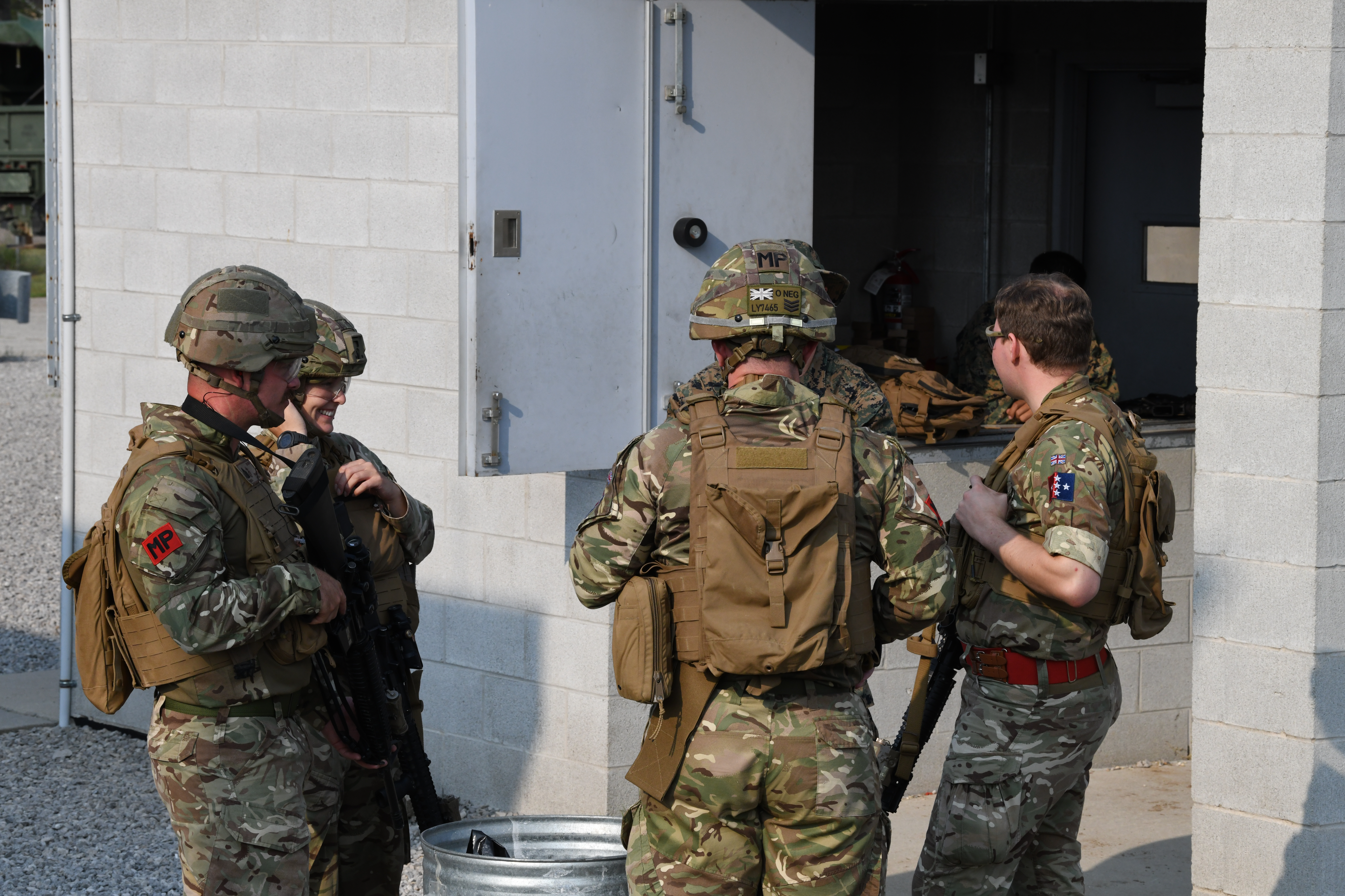 Four people in military camouflage uniforms, standing and talking near a concrete building. They are wearing helmets.