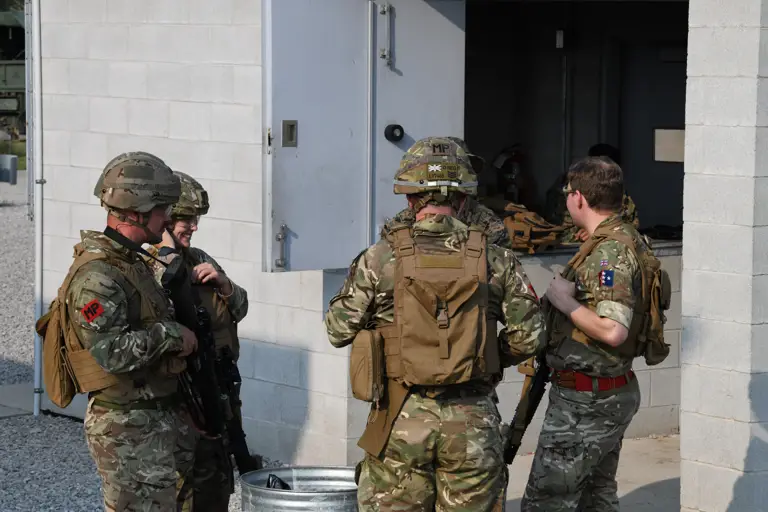 Four people in military camouflage uniforms, standing and talking near a concrete building. They are wearing helmets.