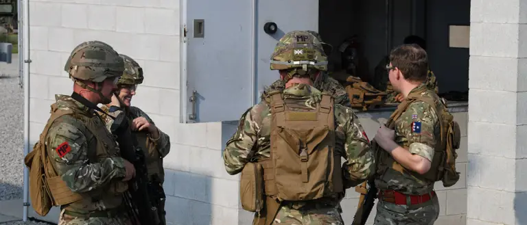Four people in military camouflage uniforms, standing and talking near a concrete building. They are wearing helmets.