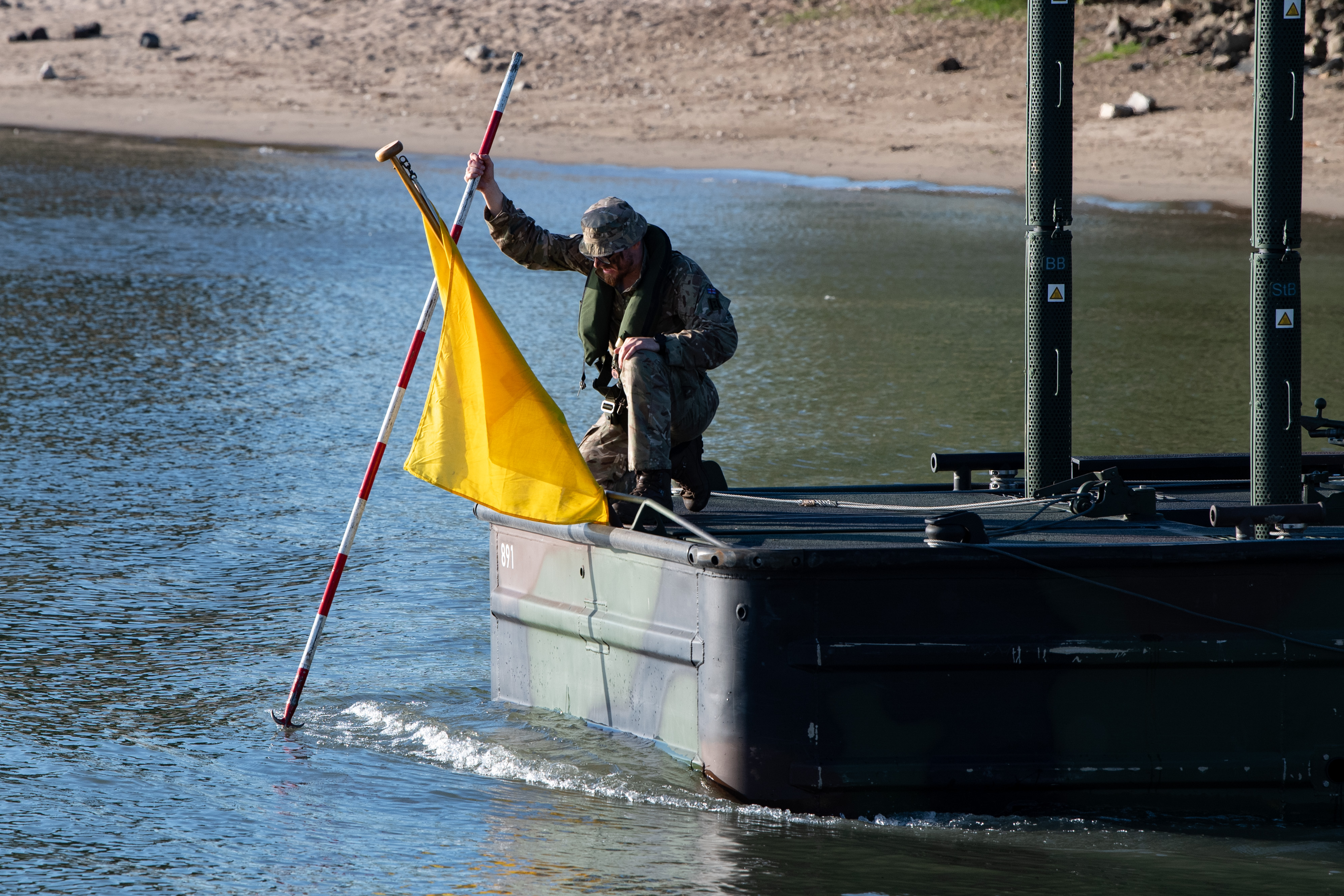 Soldier in camouflage uniform kneels on a military boat holding a yellow flag and a striped pole.