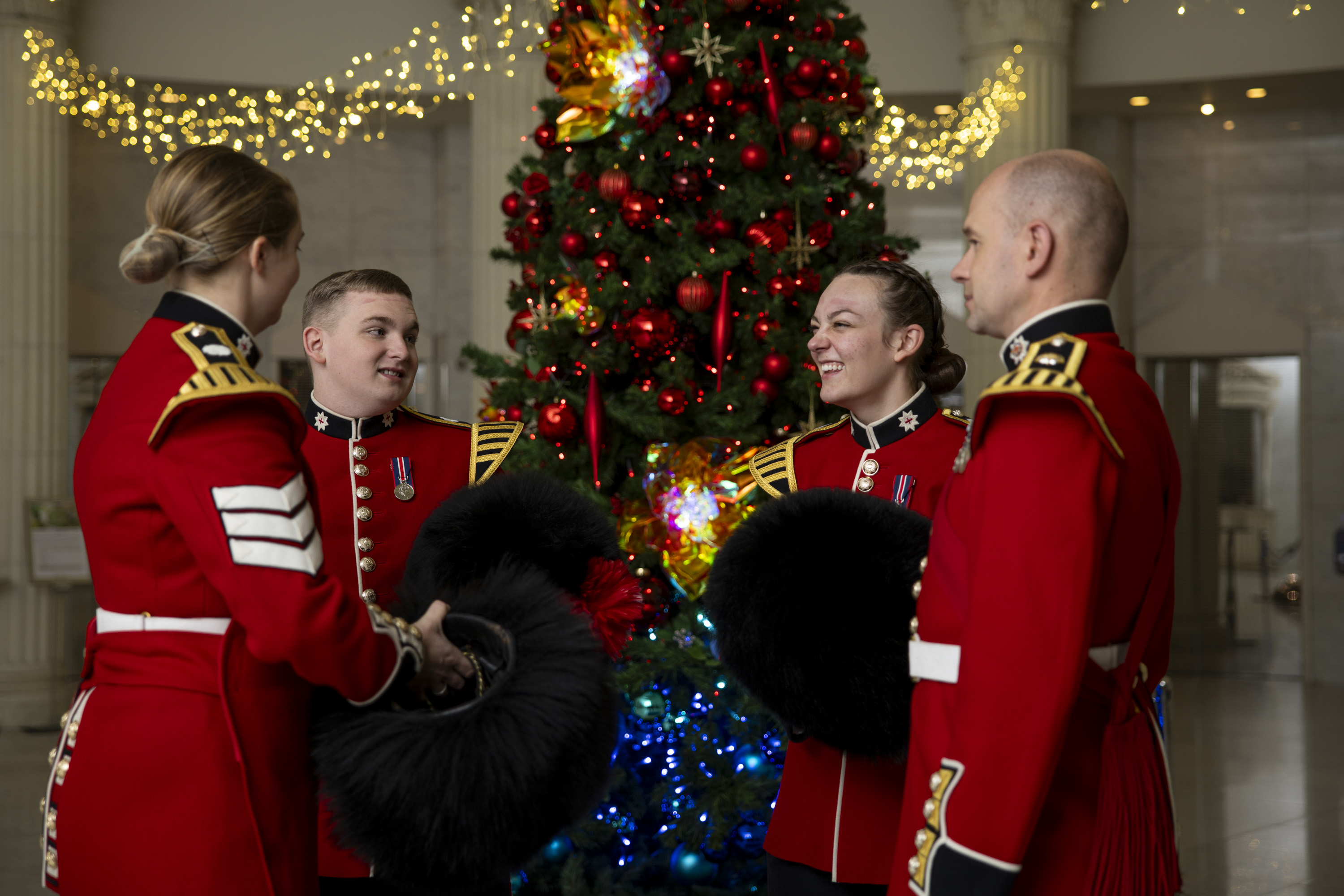Four soldiers in red tunics hold their bearskin hats and laugh next to a Christmas tree. 