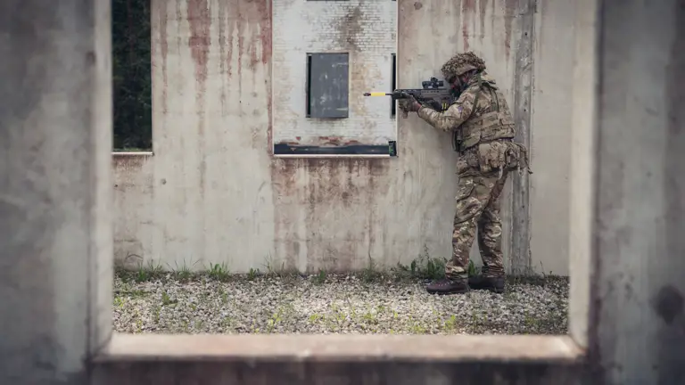 A soldier wearing camouflage uniform holds a rifle up to a window.