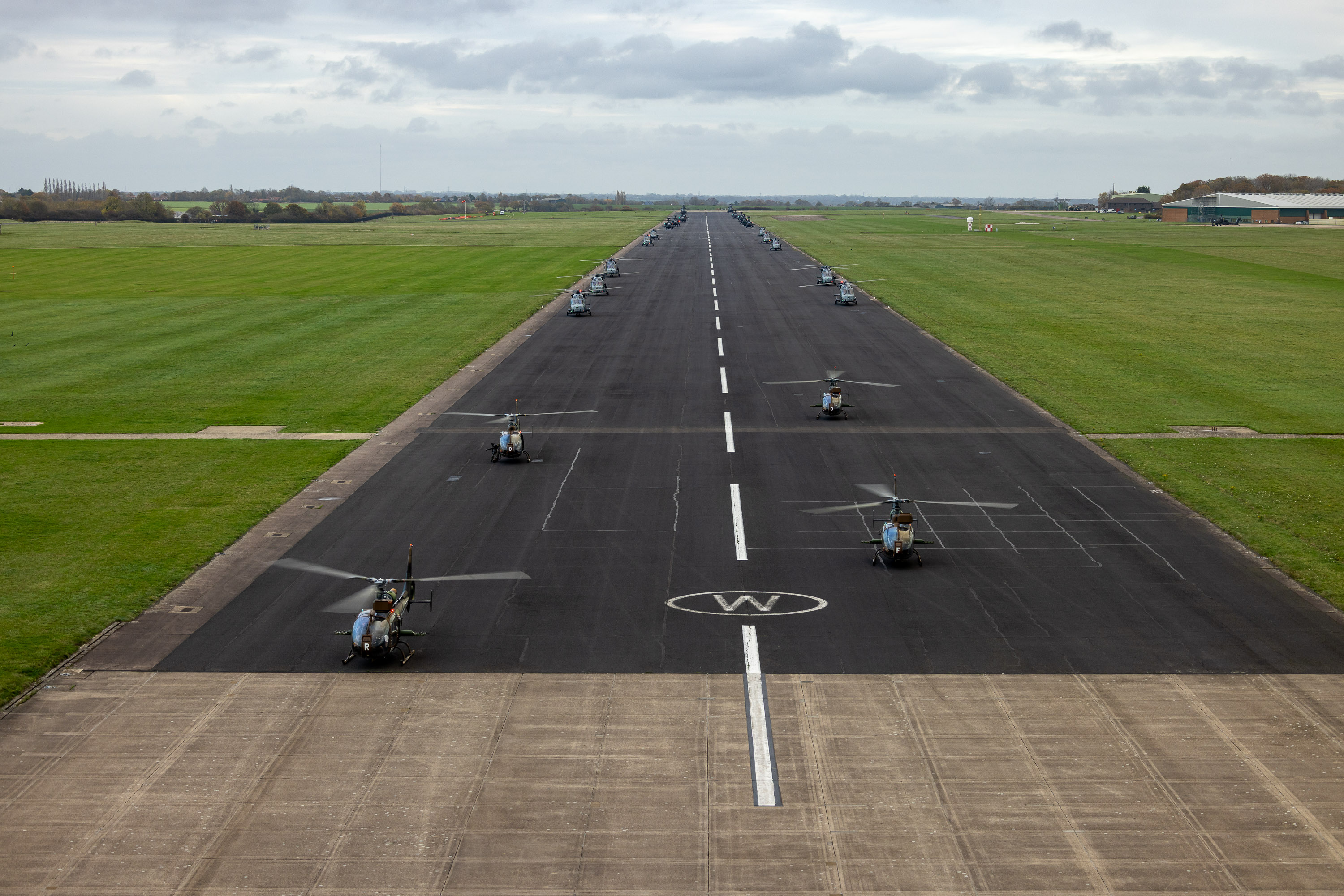 Aerial view of a runway lined with multiple helicopters parked symmetrically on both sides under a cloudy sky.