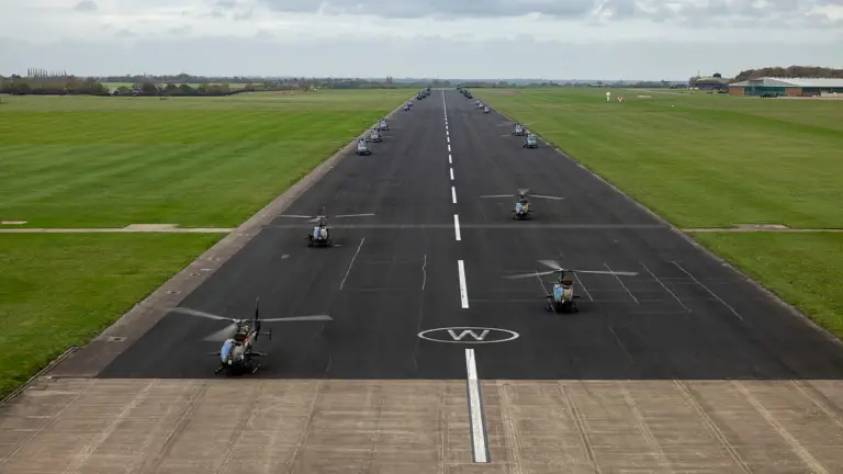Aerial view of a runway lined with multiple helicopters parked symmetrically on both sides under a cloudy sky.