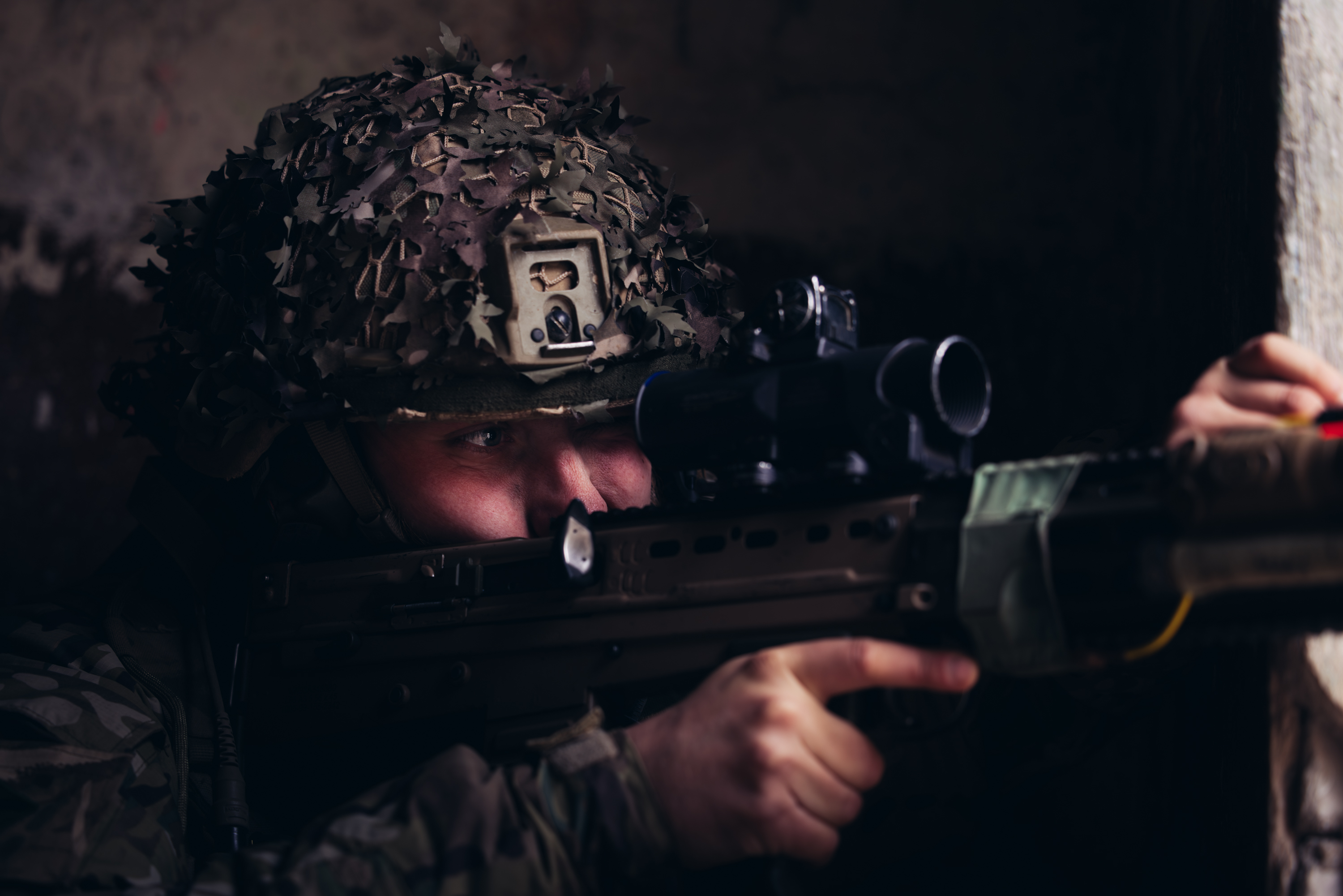 A soldier in uniform looks down the scope of their rifle.