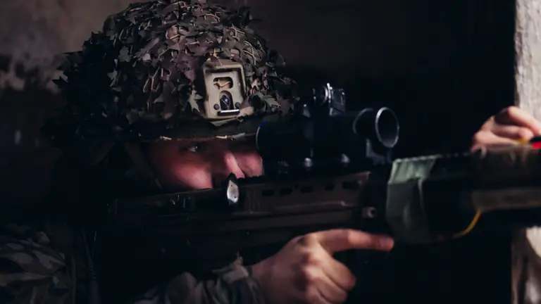 A soldier in uniform looks down the scope of their rifle.