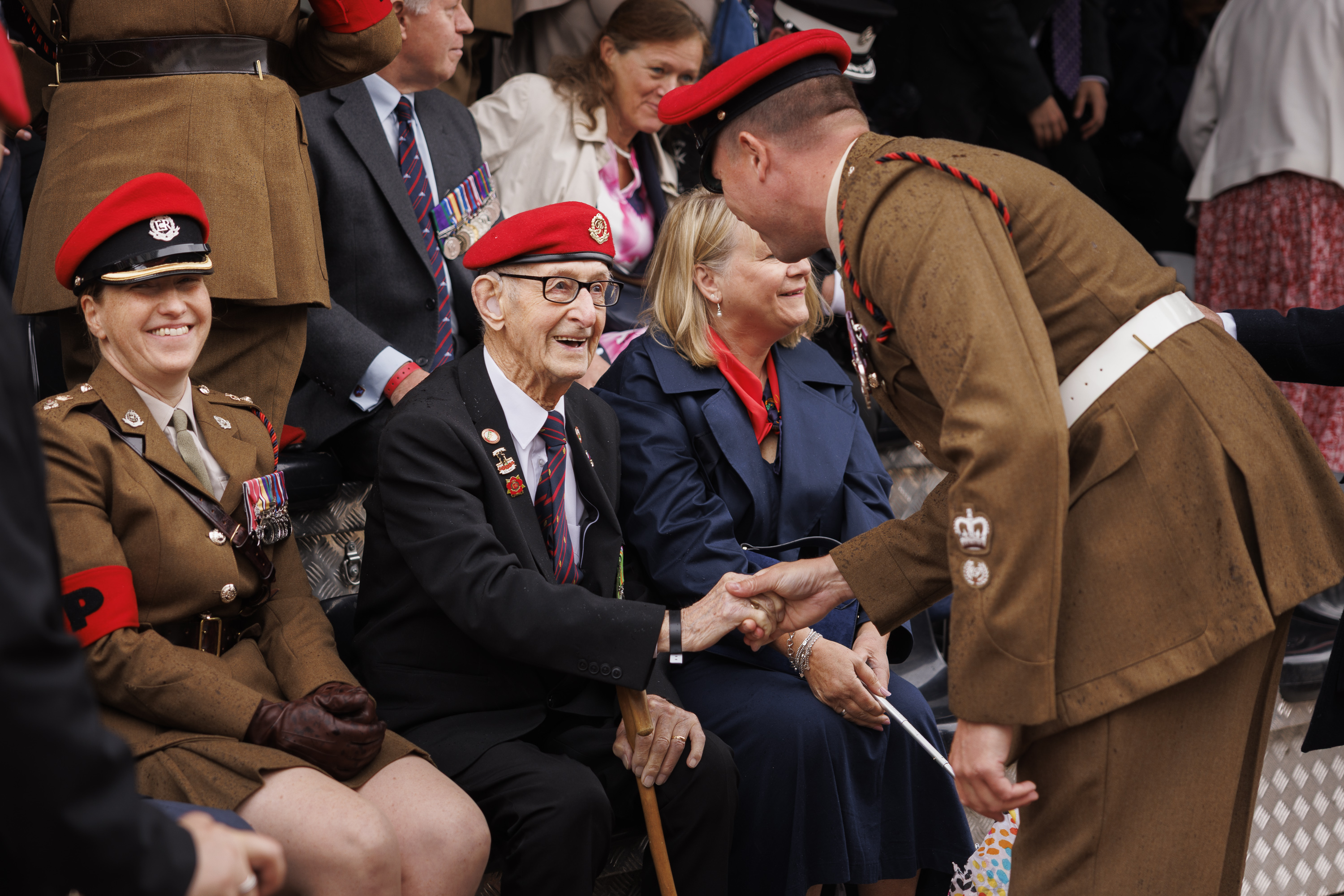 Soldiers in brown smart uniform with red caps shake hand of old man wearing a blue suit and the same red hat who is sitting