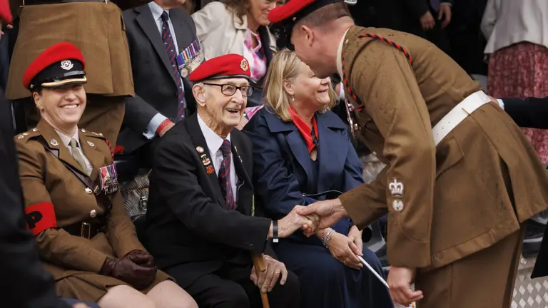 Soldiers in brown smart uniform with red caps shake hand of old man wearing a blue suit and the same red hat who is sitting
