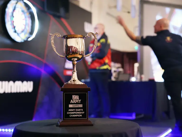 Close-up of the Army Darts Championship trophy on a table with players throwing darts in the background.
