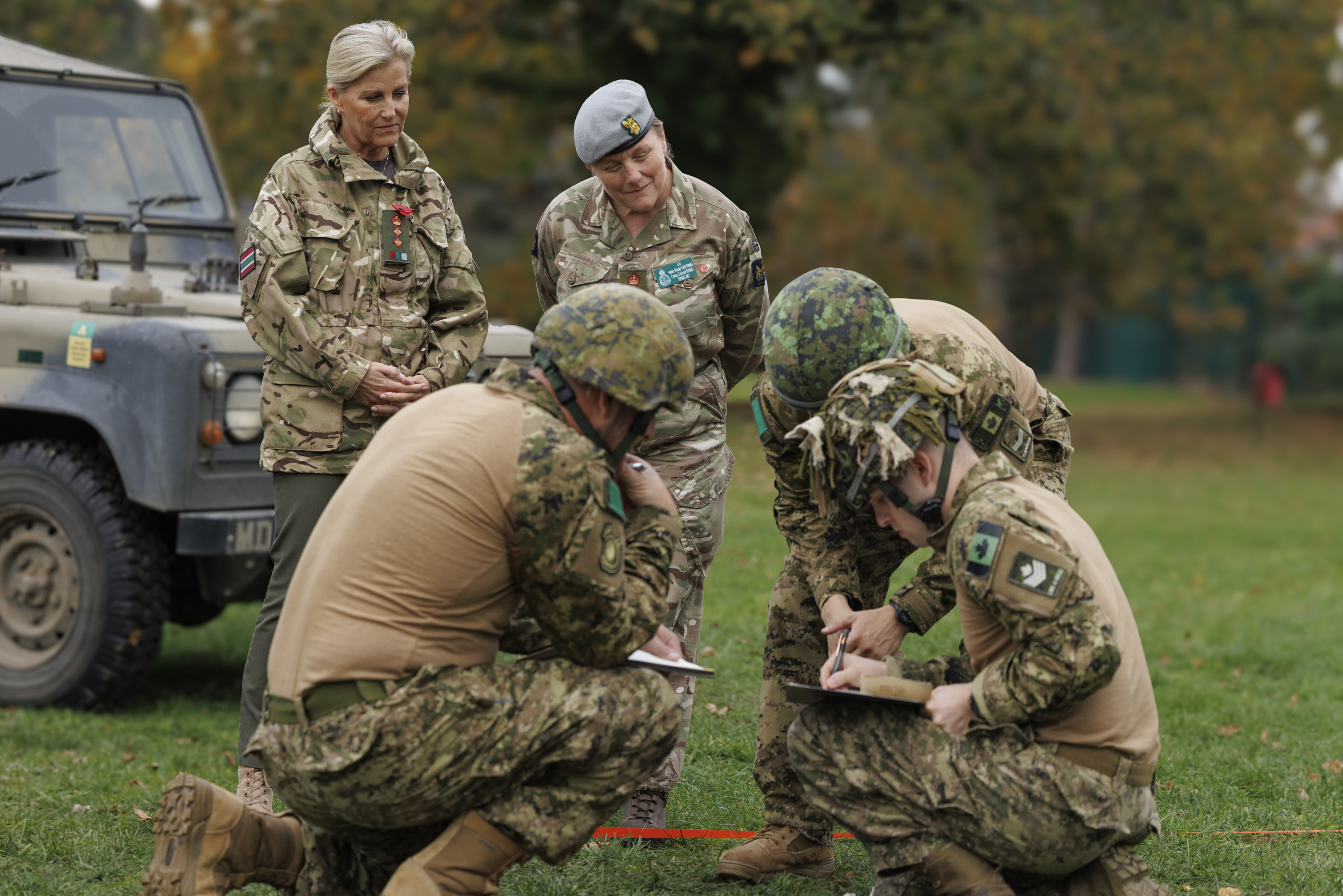 Four soldiers in camouflage uniforms and helmets are engaged in a field briefing, two others are observing. 