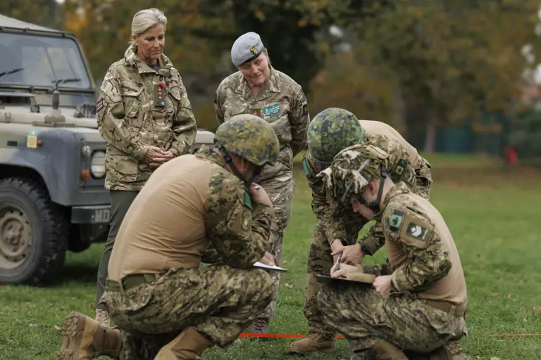 Four soldiers in camouflage uniforms and helmets are engaged in a field briefing, two others are observing. 