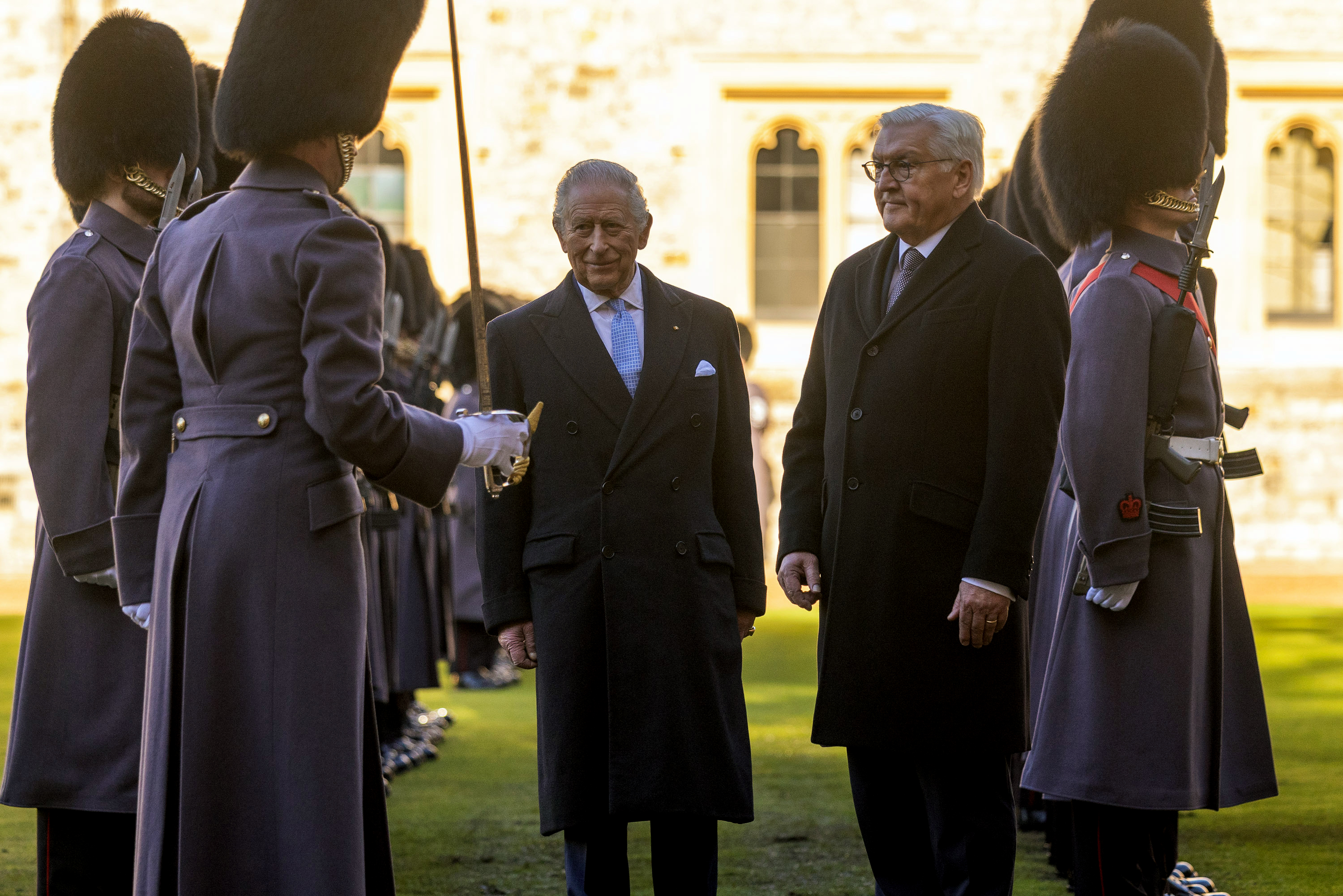 Two men in formal attire stand together, surrounded by guards in grey uniforms and bearskin hats. The setting is an outdoor ceremonial event.