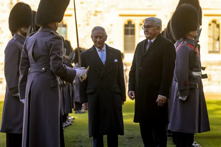 Two men in formal attire stand together, surrounded by guards in grey uniforms and bearskin hats. The setting is an outdoor ceremonial event.