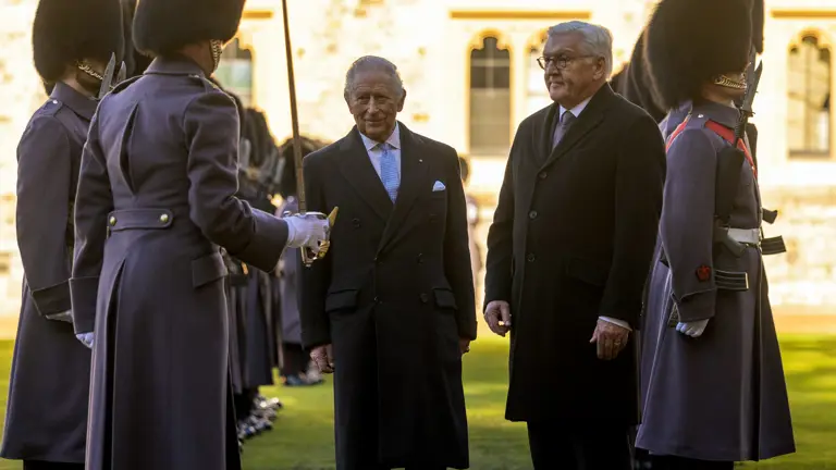 Two men in formal attire stand together, surrounded by guards in grey uniforms and bearskin hats. The setting is an outdoor ceremonial event.