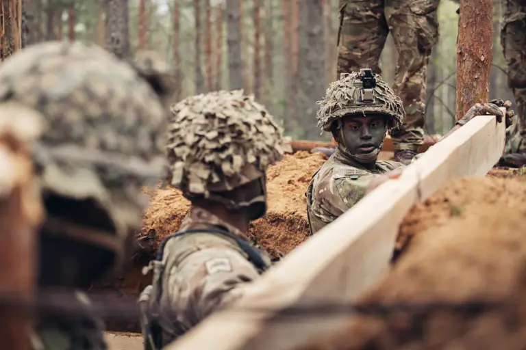 Three soldiers wearing camouflague uniforms stand in a trench. They are reinforcing the sides of the trench with wooden planks.