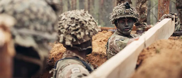 Three soldiers wearing camouflague uniforms stand in a trench. They are reinforcing the sides of the trench with wooden planks.