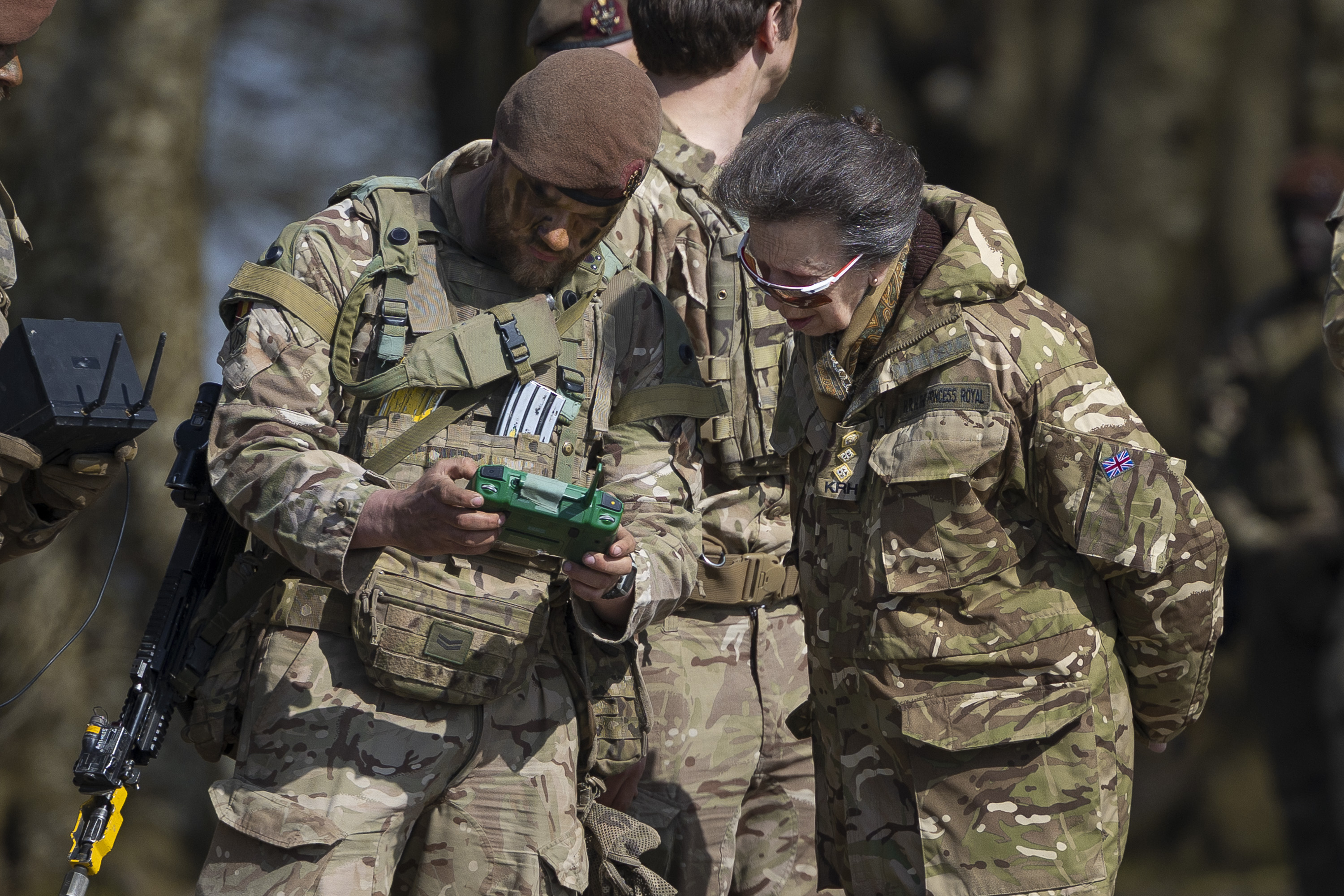 A person dressed in military camouflage shows a green device to another individual in similar attire. 