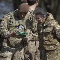 A person dressed in military camouflage shows a green device to another individual in similar attire.
