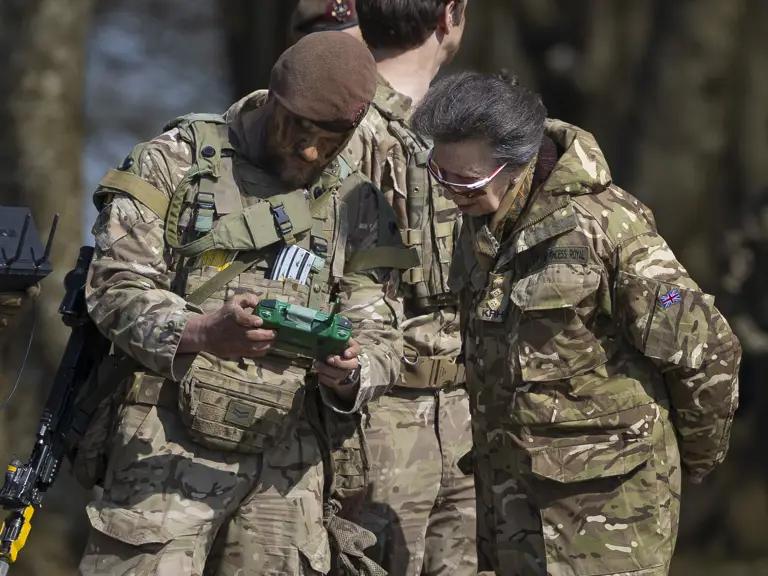 A person dressed in military camouflage shows a green device to another individual in similar attire.
