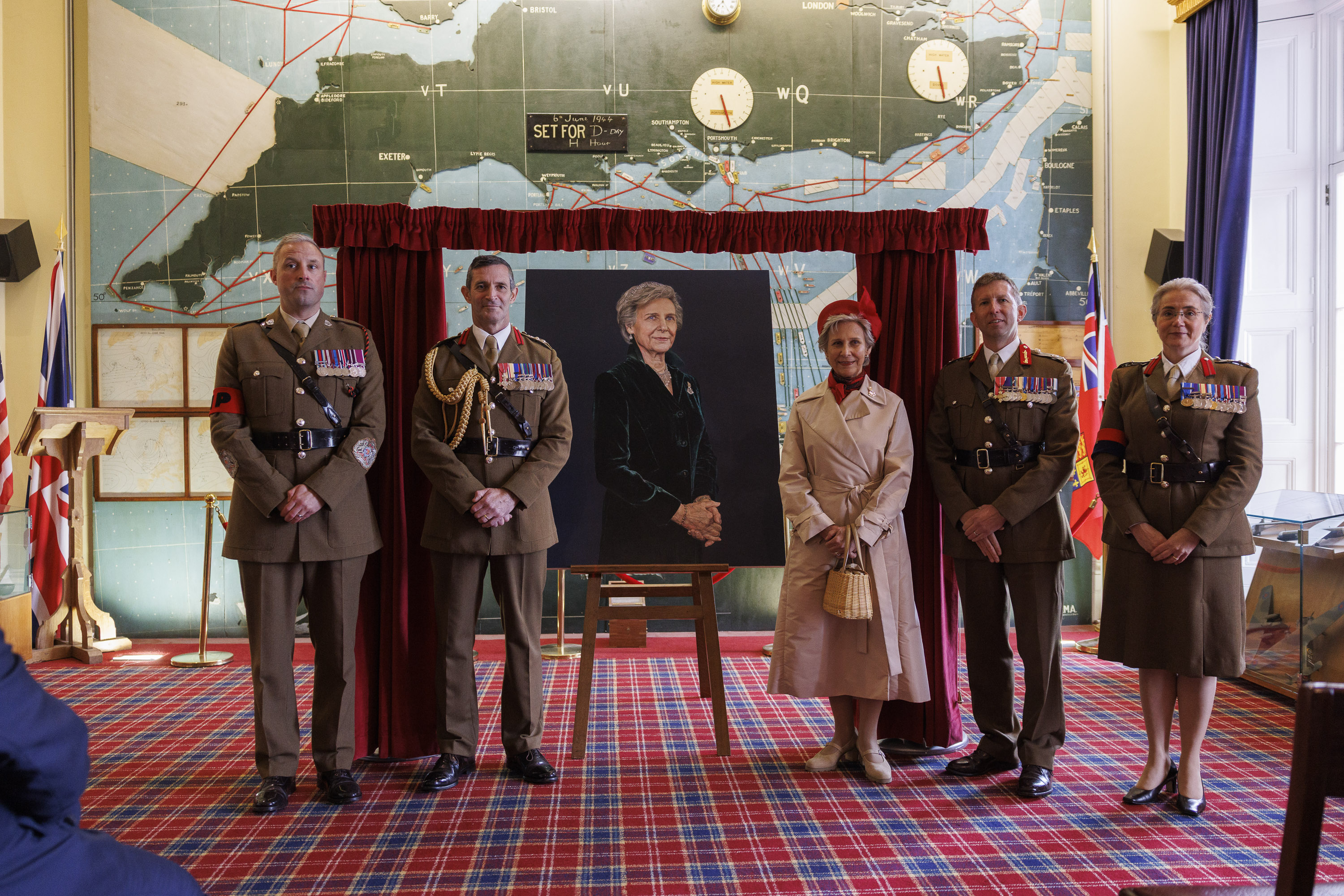 Woman in cream coat and red hat stands next to a painted portrait of her as soldiers in smart brown uniforms and no hats stand next to her.