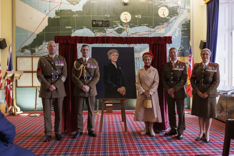 Woman in cream coat and red hat stands next to a painted portrait of her as soldiers in smart brown uniforms and no hats stand next to her.
