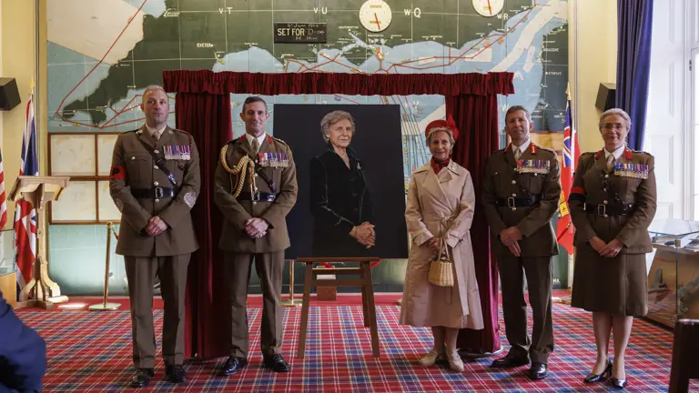 Woman in cream coat and red hat stands next to a painted portrait of her as soldiers in smart brown uniforms and no hats stand next to her.