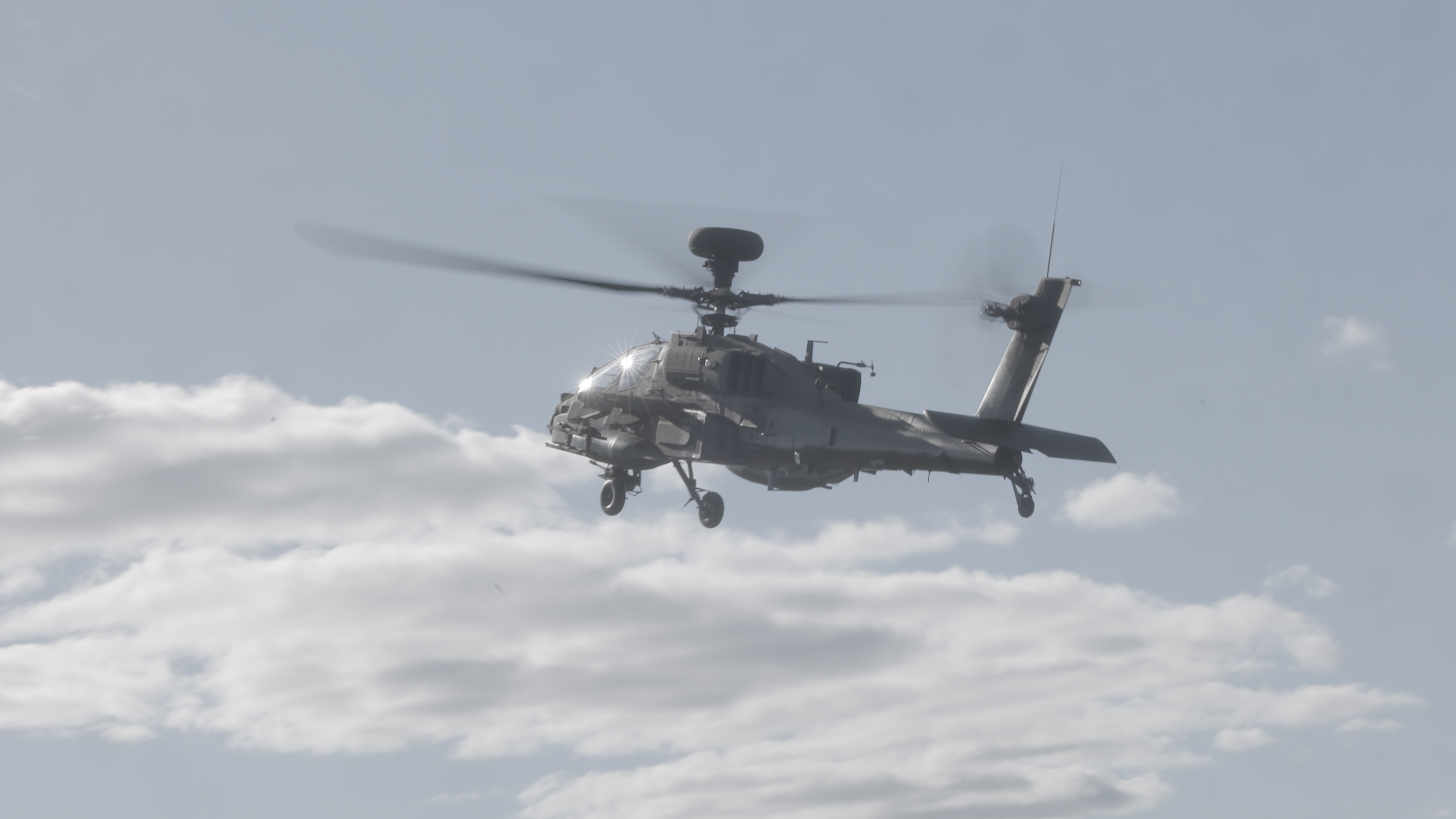 Image shows a AH-64E Apache helicopter mid flight in the sky. 