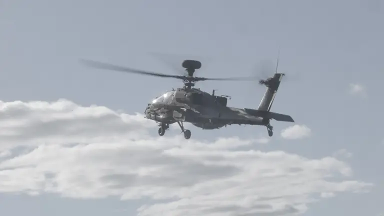 Image shows a AH-64E Apache helicopter mid flight in the sky.