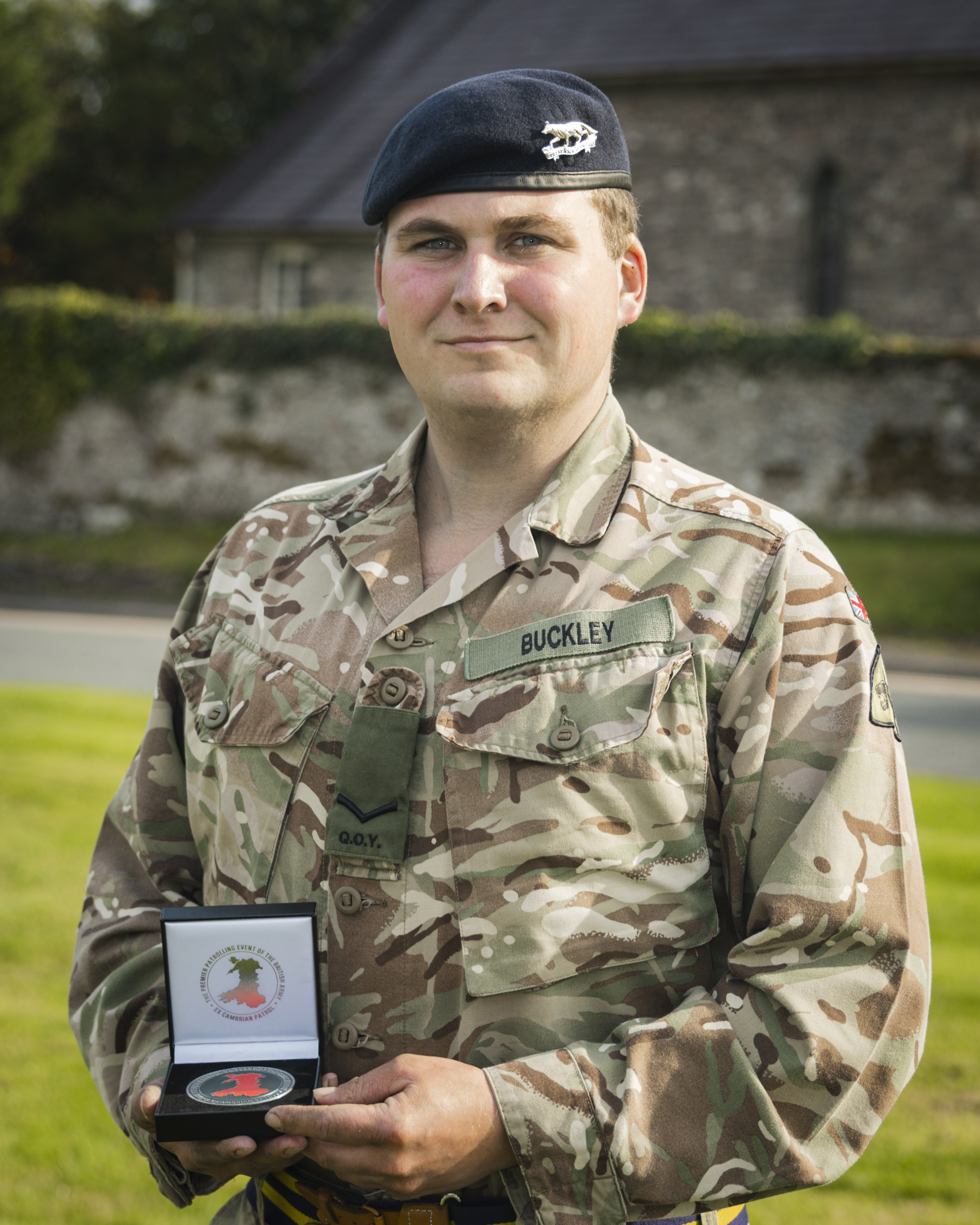 Soldier in camouflage uniform and beret holding an open medal case outdoors near a stone building and greenery.