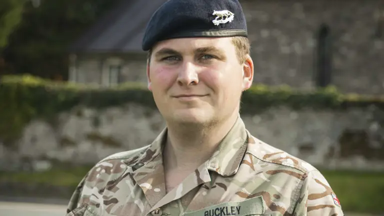 Soldier in camouflage uniform and beret holding an open medal case outdoors near a stone building and greenery.