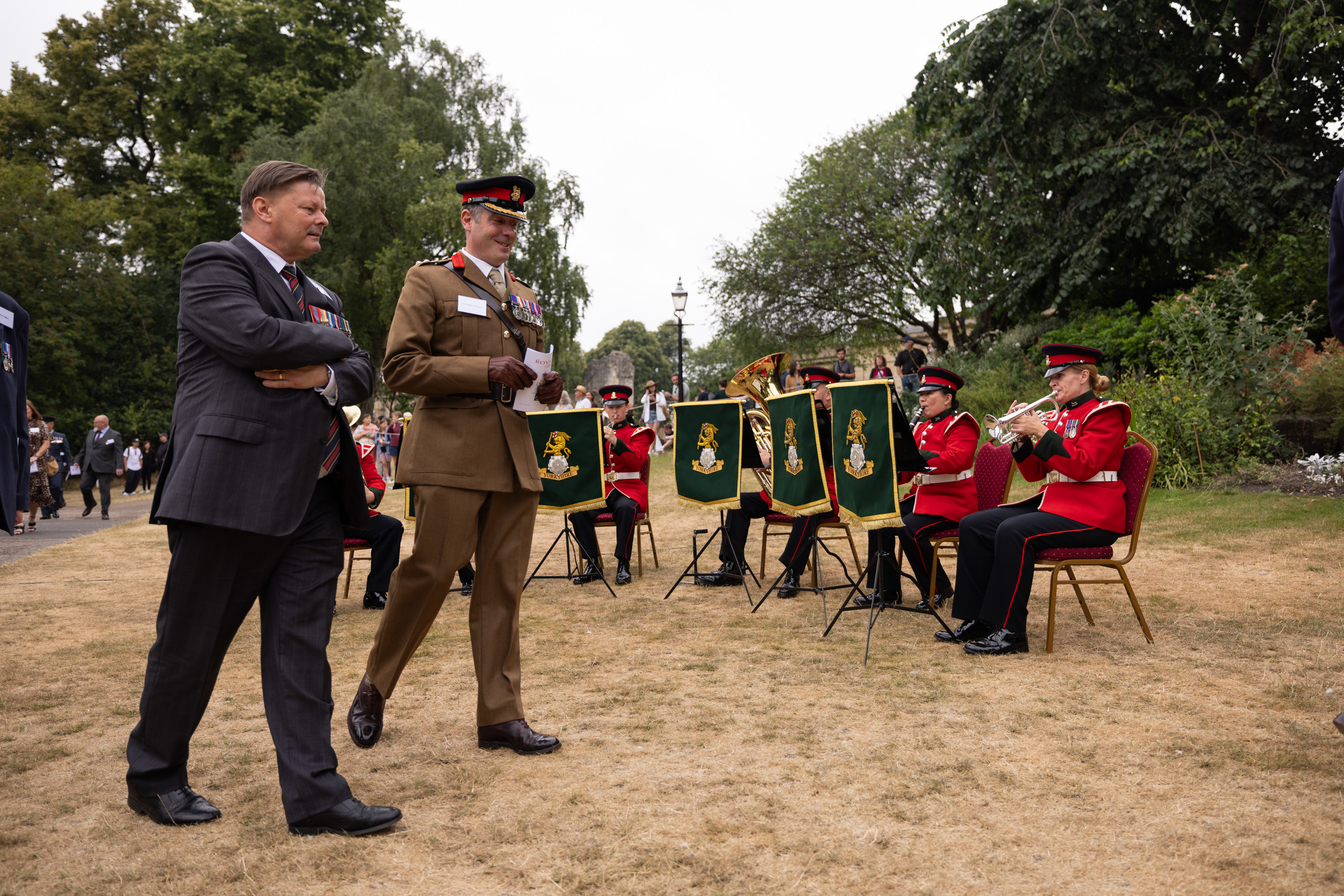 A military band dressed in red uniforms is playing music outdoors. Two individuals are walking past.
