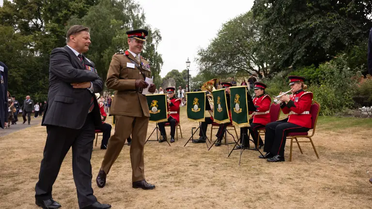 A military band dressed in red uniforms is playing music outdoors. Two individuals are walking past.