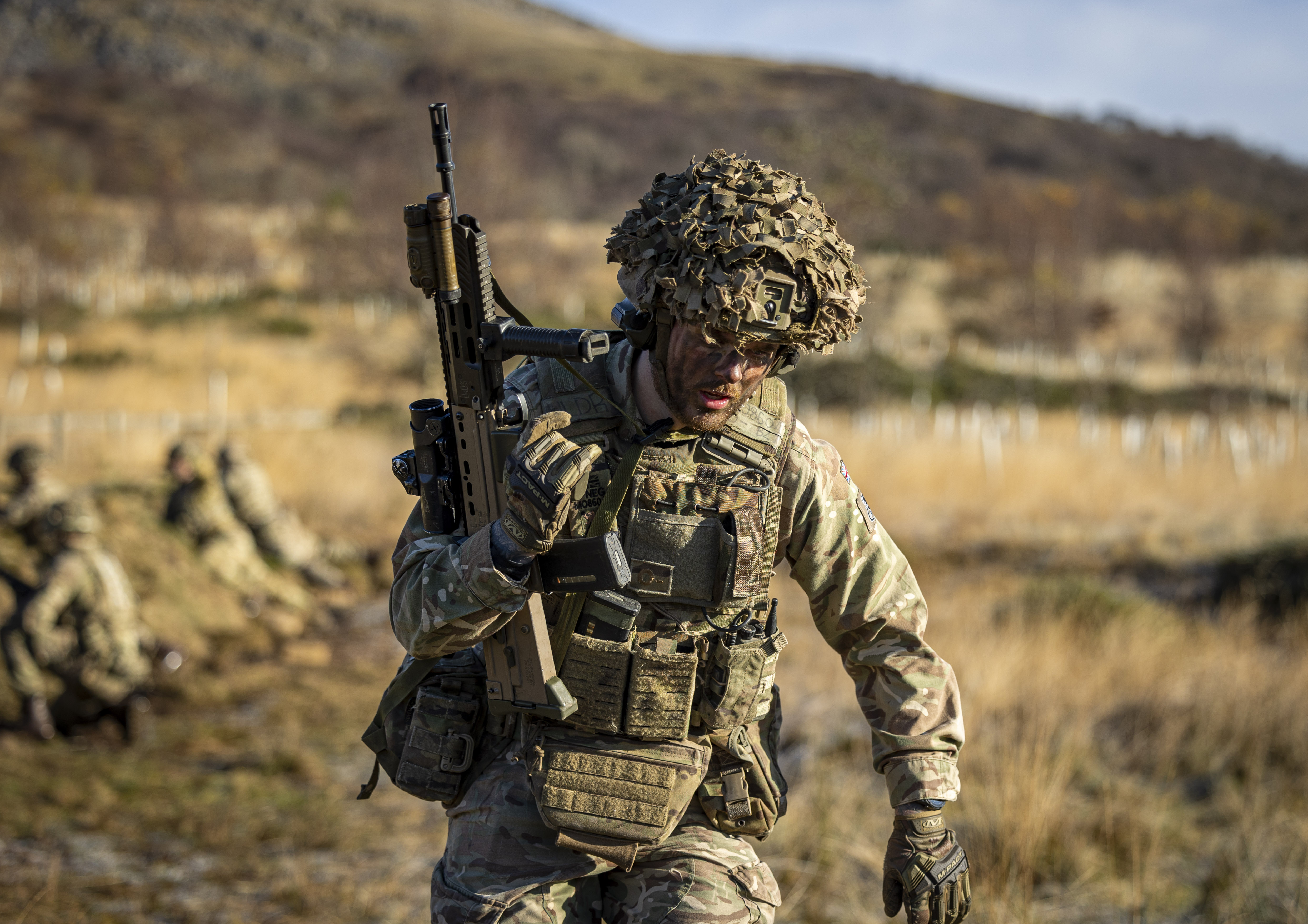 A man in uniform is pictured holding a weapon upright in a grassy landscape. 