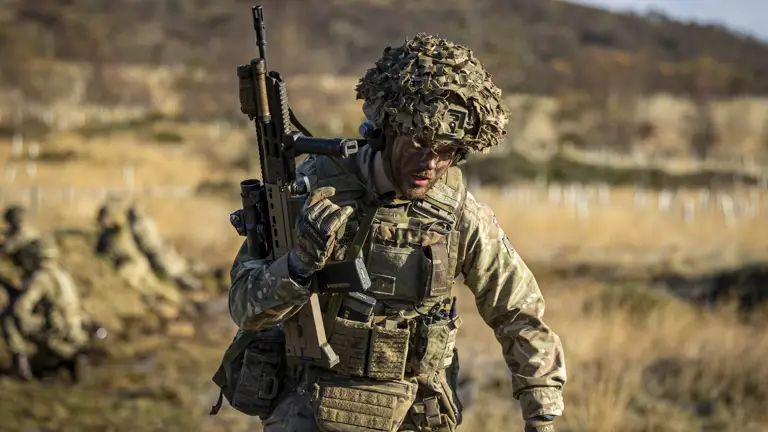 A man in uniform is pictured holding a weapon upright in a grassy landscape.