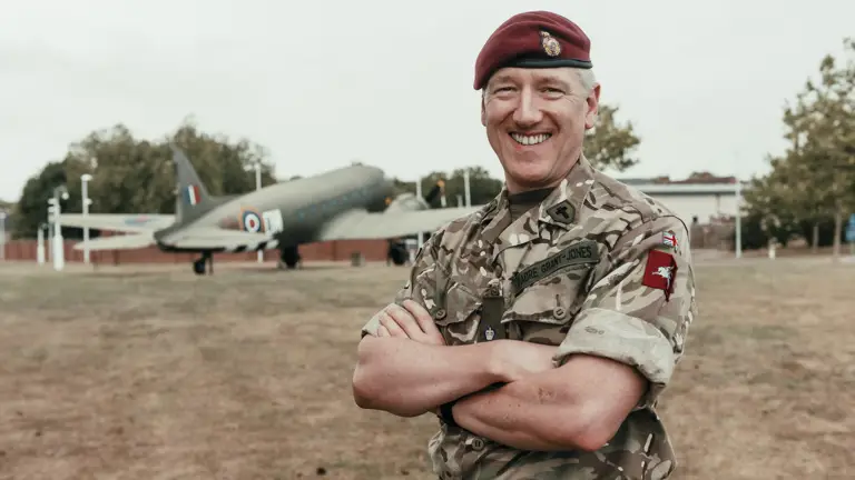 Reverend Mark Grant-Jones is posing for a portrait stood in front of a Gate Guardian Dakota Aircraft. He is wearing this camouflage uniform and beret.