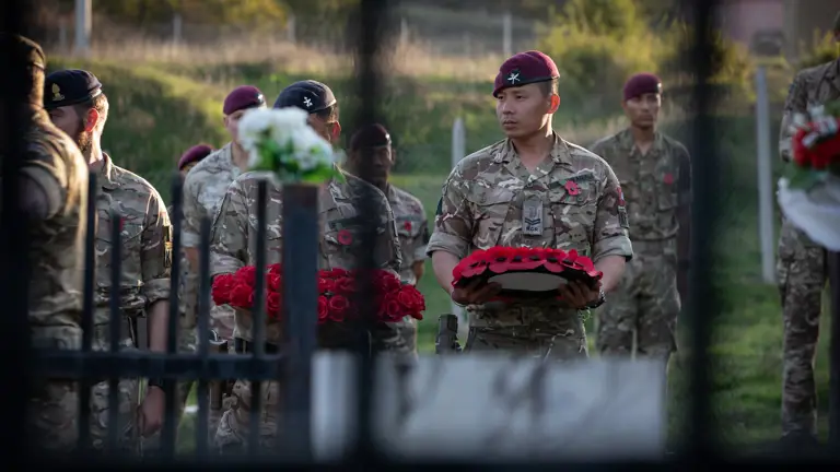 A soldier wearing camouflage uniform and a maroon beret holds a poppy wreath.