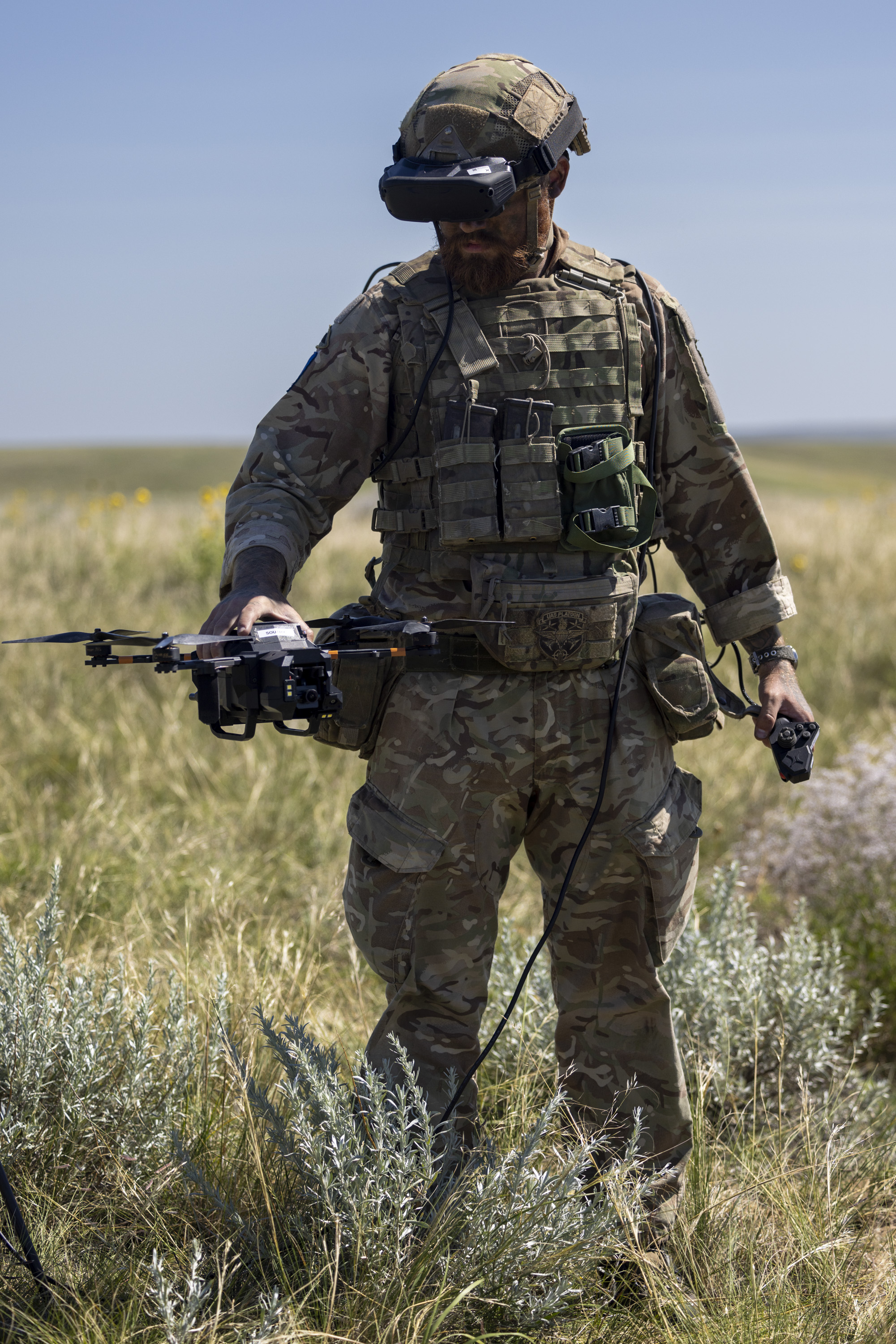 A person in camouflage military gear stands in an open grassy field, holding a drone. The person is wearing a helmet and protective eyewear, with various equipment attached to their vest. The sky is clear and blue.