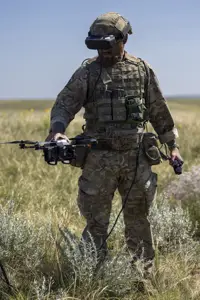 A person in camouflage military gear stands in an open grassy field, holding a drone. The person is wearing a helmet and protective eyewear, with various equipment attached to their vest. The sky is clear and blue.