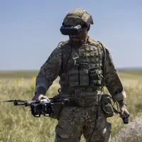 A person in camouflage military gear stands in an open grassy field, holding a drone. The person is wearing a helmet and protective eyewear, with various equipment attached to their vest. The sky is clear and blue.