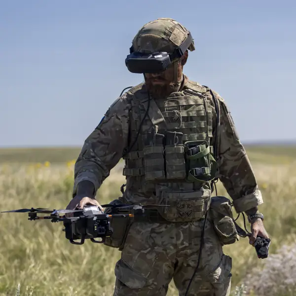 A person in camouflage military gear stands in an open grassy field, holding a drone. The person is wearing a helmet and protective eyewear, with various equipment attached to their vest. The sky is clear and blue.