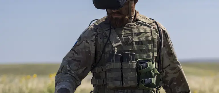 A person in camouflage military gear stands in an open grassy field, holding a drone. The person is wearing a helmet and protective eyewear, with various equipment attached to their vest. The sky is clear and blue.
